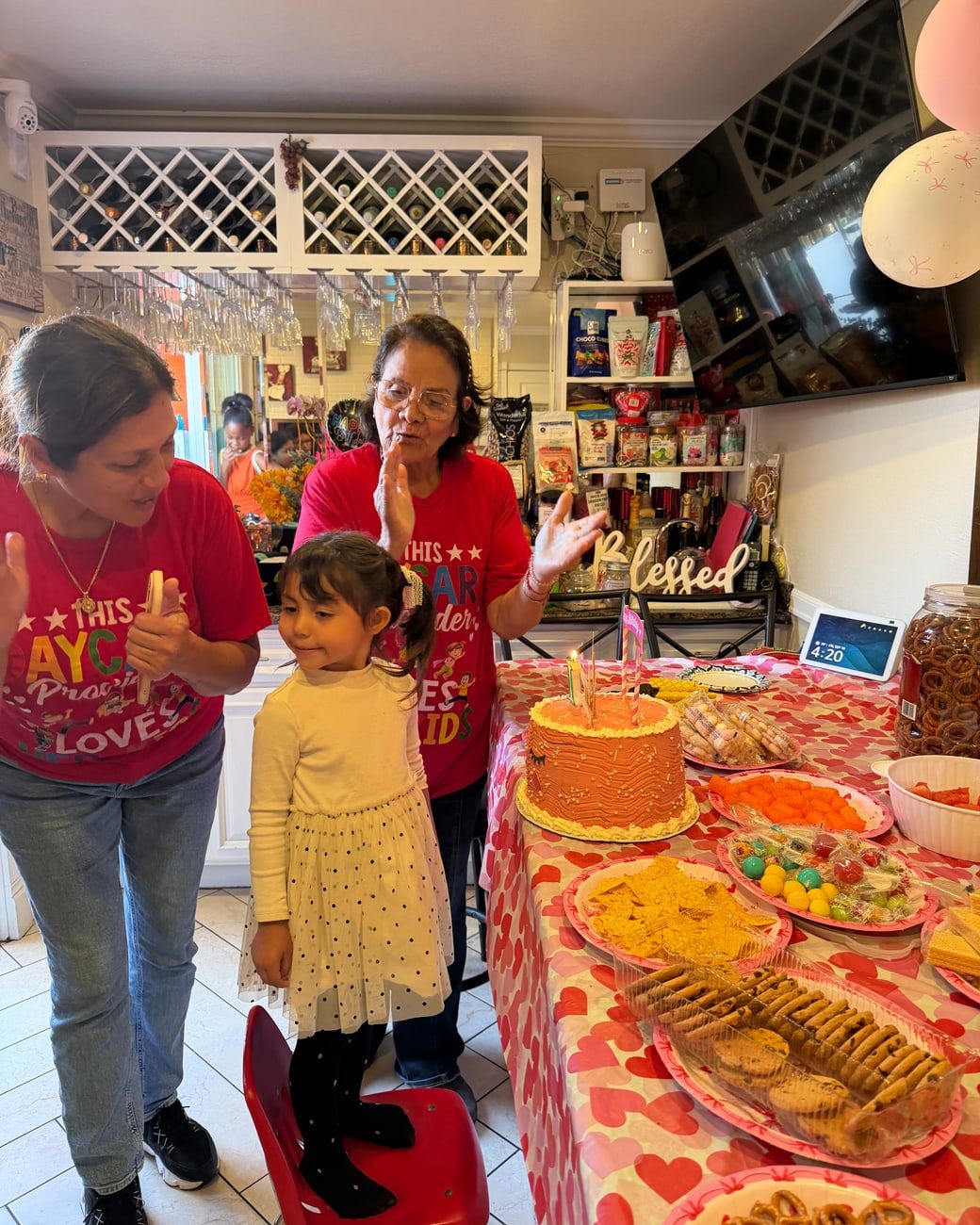 Two caregivers in red daycare shirts cheering as a girl gets ready to blow out her birthday candles