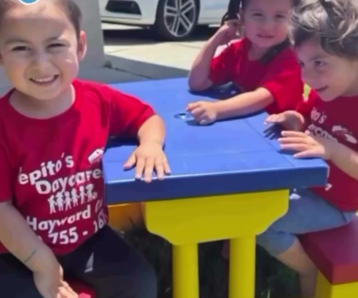 Three kids smiling at a blue and yellow picnic table